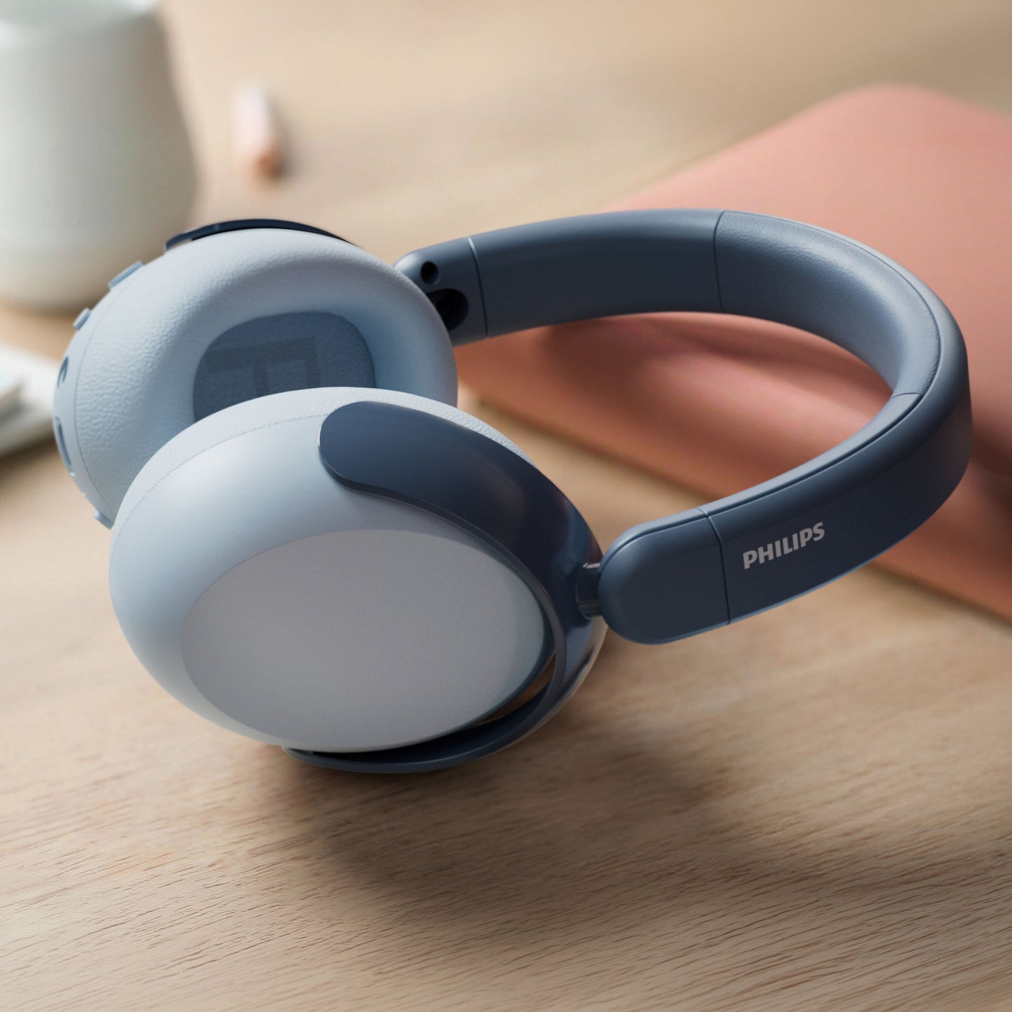 Philips 5000-serien trådløse blå over-ear børnehovedtelefoner på træbord, sløret ferskenfarvet objekt og hvidt krus bagved.