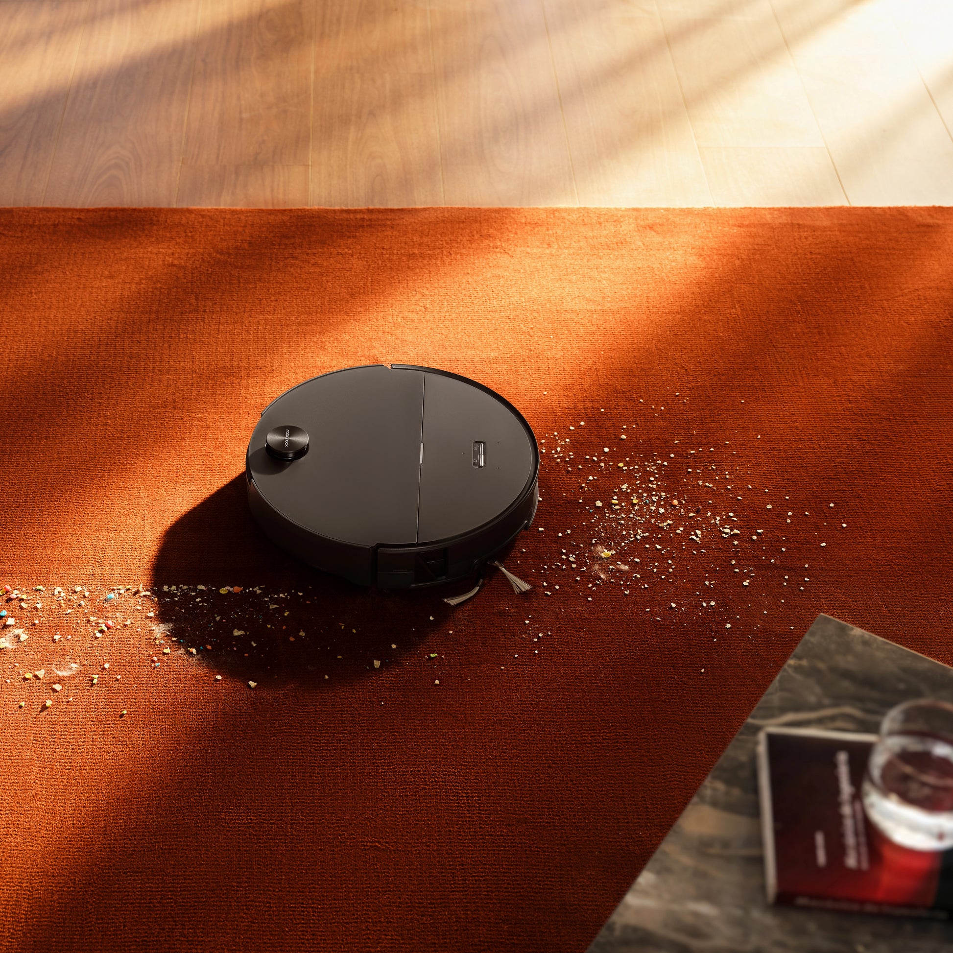 Roborock Saros 20 Sonic robotstøvsuger med vibrerende moppe på orange tæppe, glas og bog synlige på bord ved siden af.