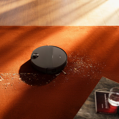 Roborock Saros 20 Sonic robotstøvsuger med vibrerende moppe på orange tæppe, glas og bog synlige på bord ved siden af.