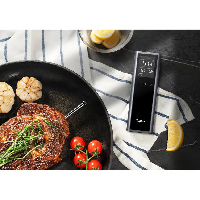 Typur Sync One trådløst stegetermometer med smart basestation viser temperaturen, placeret ved siden af panden med bøffen og tilbehøret.