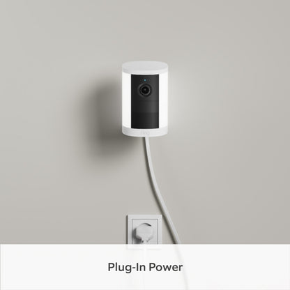 Ring Spotlight Cam Plug-in Retinal 2K, monteret på en lysegrå væg, tilsluttet det nederste stik med et hvidt ledning.