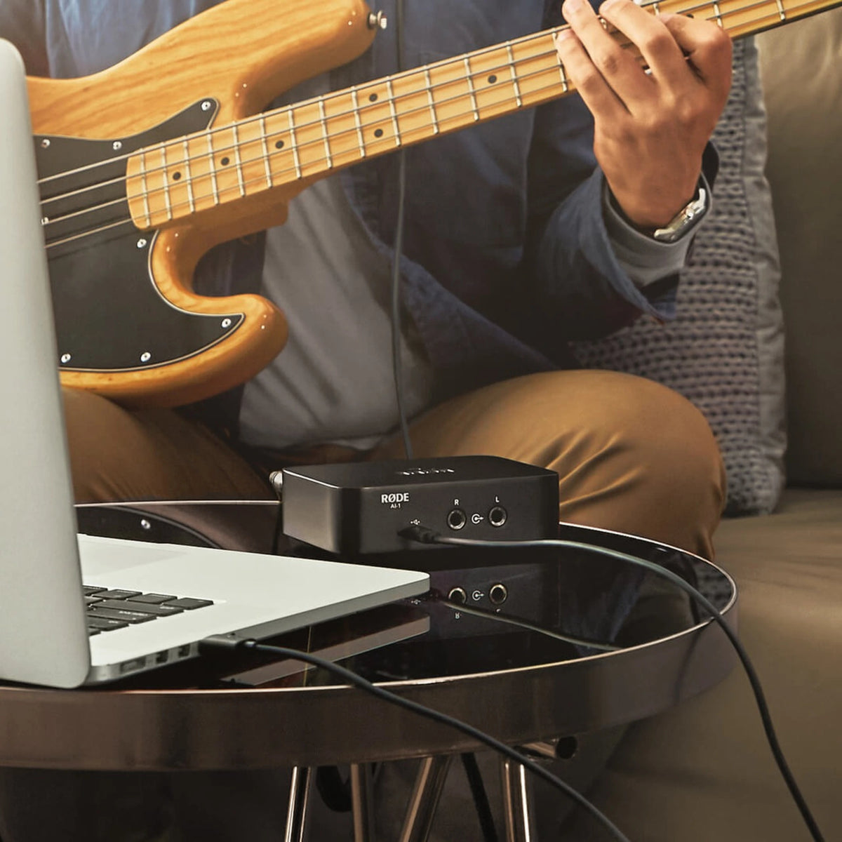 RØDE AI-1 USB-lydinterface i sort på et rundt glasbord med bærbar computer, synlige kabler, elbas og hjemmestudieopsætning.