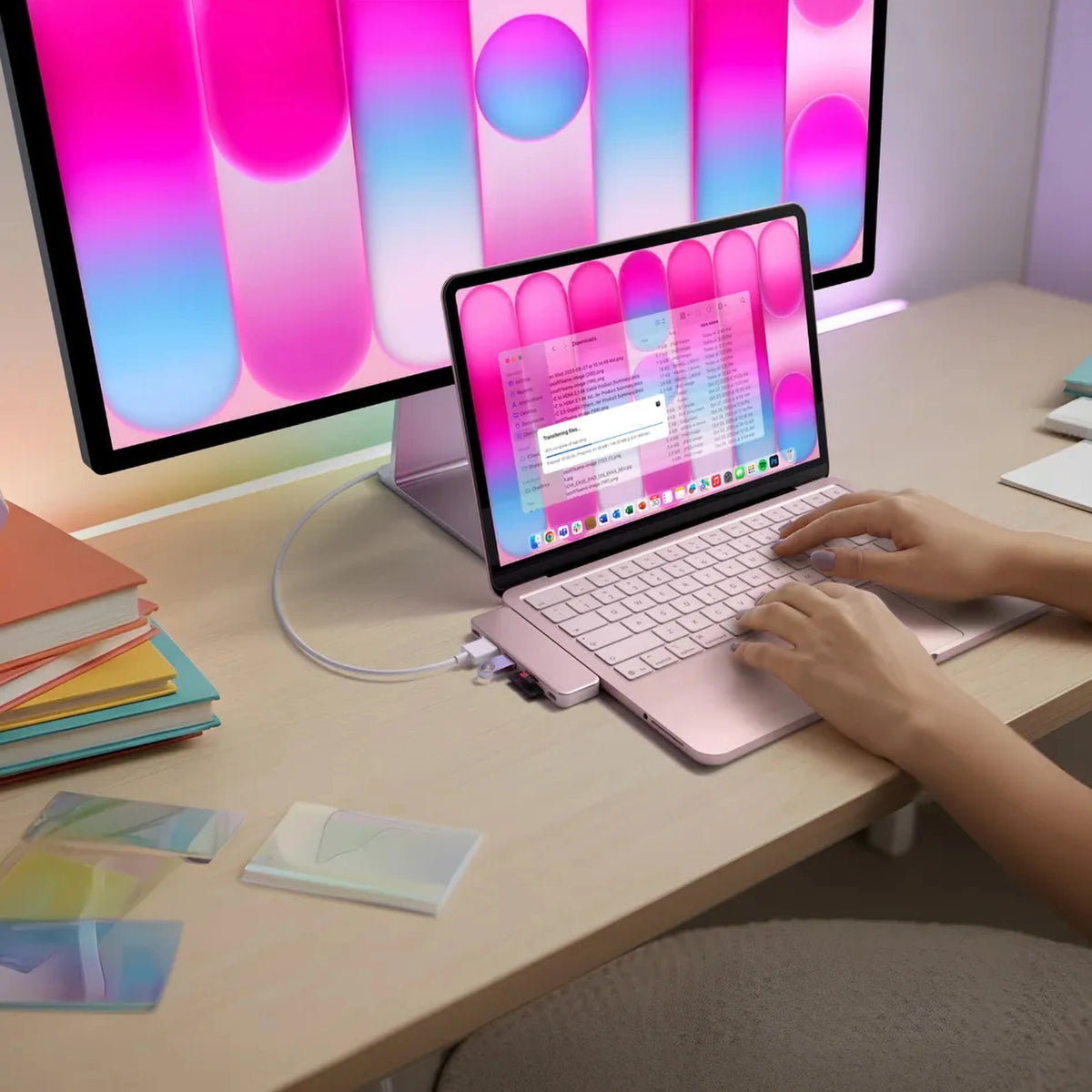 En lyserød MacBook Neo med en Satechi USB-C Snap Hub Neo Rouge tilsluttet en ekstern skærm, bøger og kort på et træbord.
