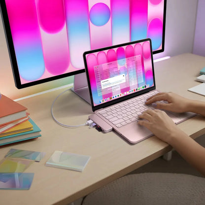 En lyserød MacBook Neo med en Satechi USB-C Snap Hub Neo Rouge tilsluttet en ekstern skærm, bøger og kort på et træbord.