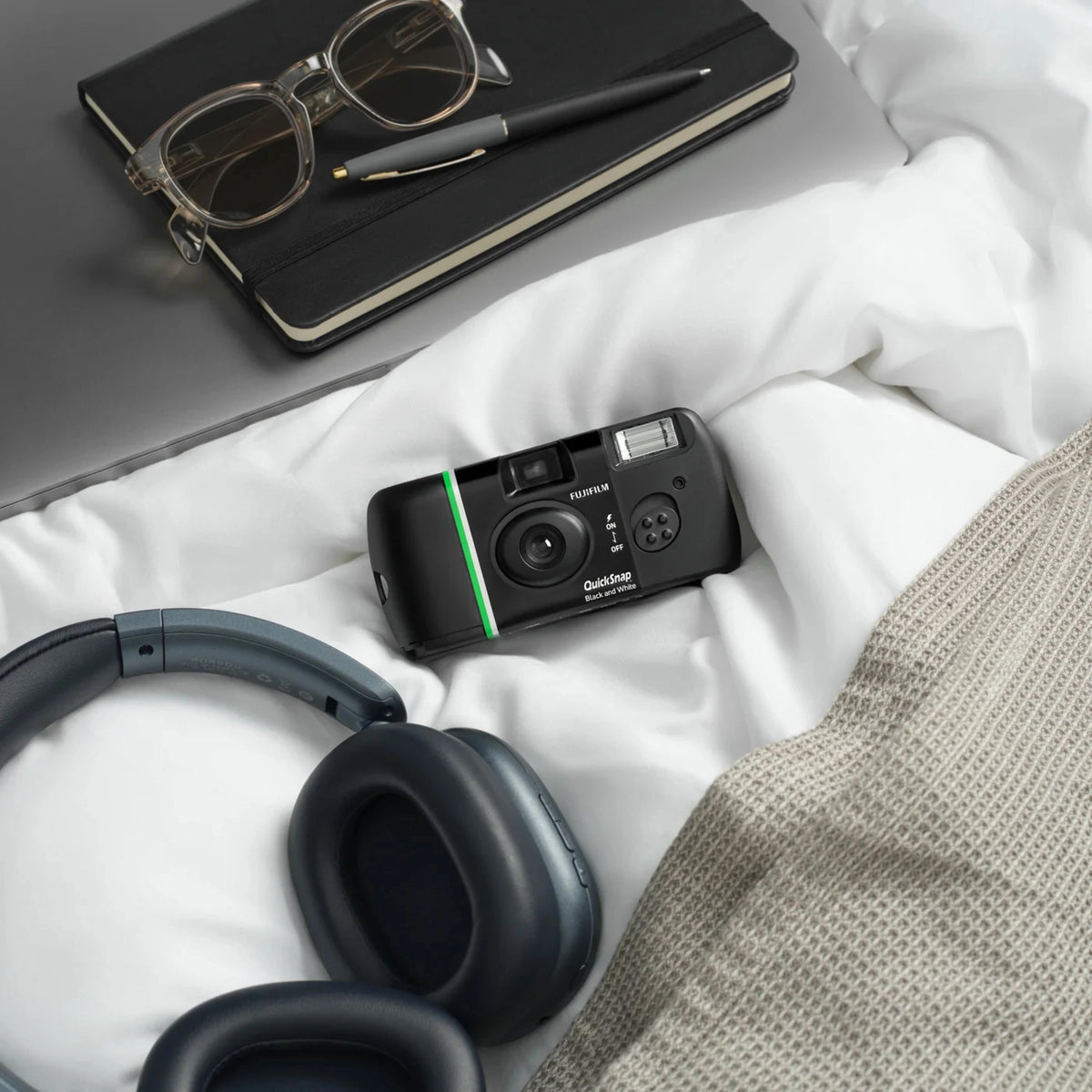 Fujifilm QuickSnap sort-hvidt engangskamera til 27 billeder, trådløse hovedtelefoner, gråt strikket tæppe, sort notesbog.