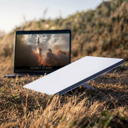 Starlink Mini Kit-satellitterminal på stativ i tørt græs ved siden af en bærbar computer med billede af en raketopsendelse.