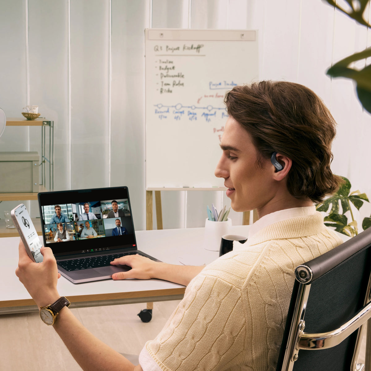 Shokz OpenFit Air 2-høretelefoner med åbne øreklapper i sort bæres af en mand under et videomøde på en bærbar computer med en smartphone.