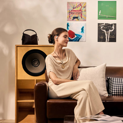 Shokz OpenFit Air 2-høretelefoner med åbne ørepropper i hvidt, båret af en kvinde, set fra siden på en brun lædersofa.
