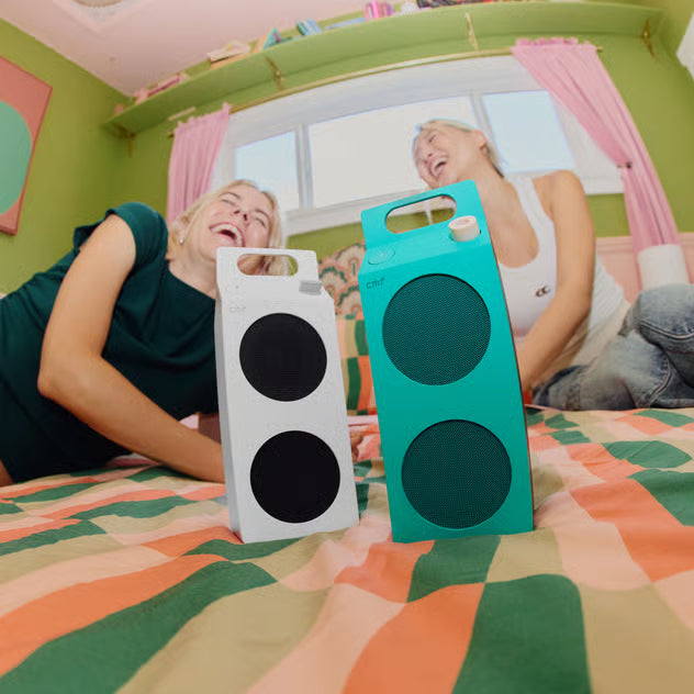 Nothing CMF Speaker M – bærbar Bluetooth-højttaler i hvid og turkis med håndtag, placeret på en seng.