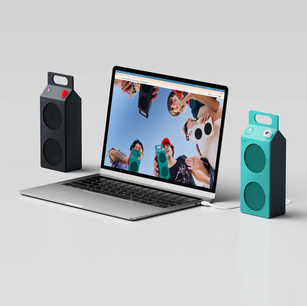 Turkis Nothing CMF Speaker M Bluetooth 6.0-højttalere med bærehåndtag ved siden af en åben bærbar computer med gruppebillede.