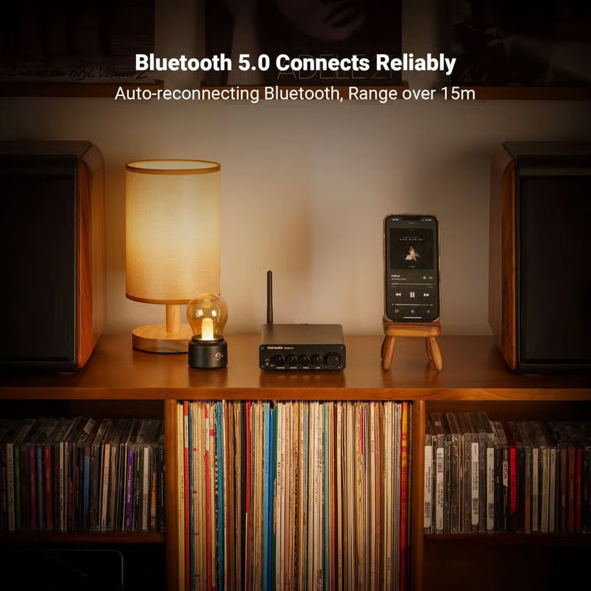 Fosi Audio BT30D Pro 2.1-forstærker med Bluetooth, 48 V og ekstern antenne placeret på en træhylde ved siden af lampen og smartphonen.