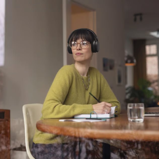 En kvinde med sorte, trådløse Philips 5000 Series over-ear-hovedtelefoner sidder ved et bord og skriver i en notesbog.