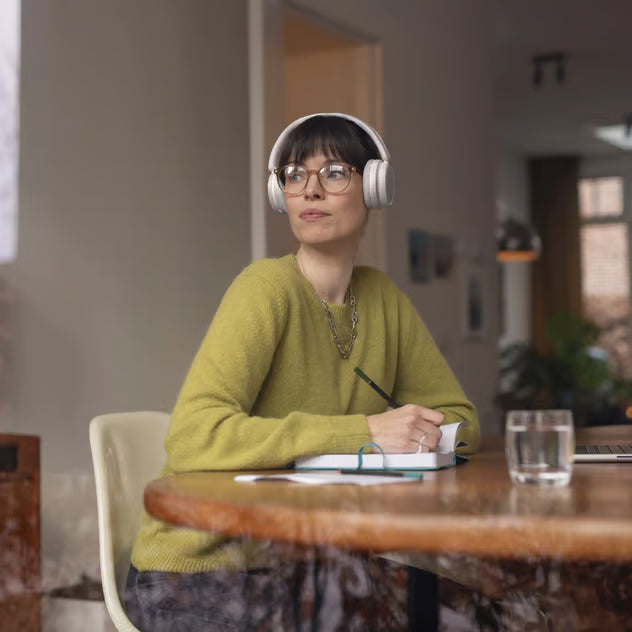 Hvide trådløse Philips 5000-serie over-ear-hovedtelefoner på en kvinde ved et bord med en notesbog og en pen.