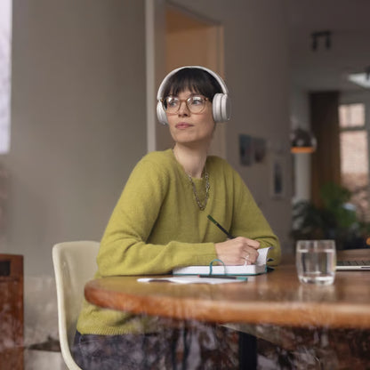 Hvide trådløse Philips 5000-serie over-ear-hovedtelefoner på en kvinde ved et bord med en notesbog og en pen.