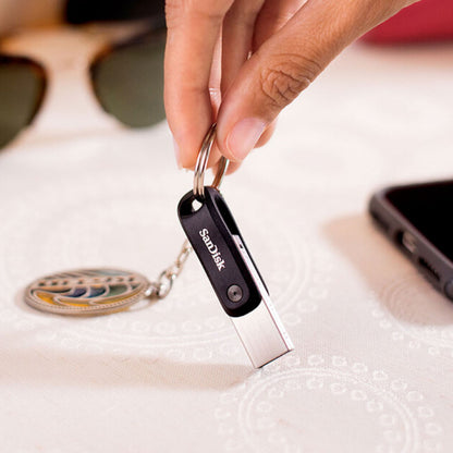 Sandisk iXpand Go USB-nøgle 256 GB med Lightning-stik, holder i hånd, smartphone og solbriller i baggrunden.