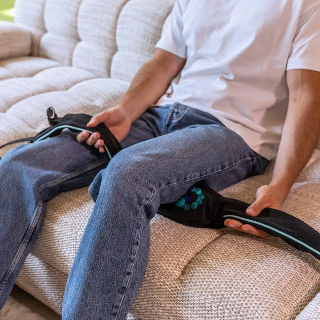 En person bruger en sort Medivon Motion X trådløs Shiatsu-nakkemassager på låret, mens vedkommende sidder i sofaen.