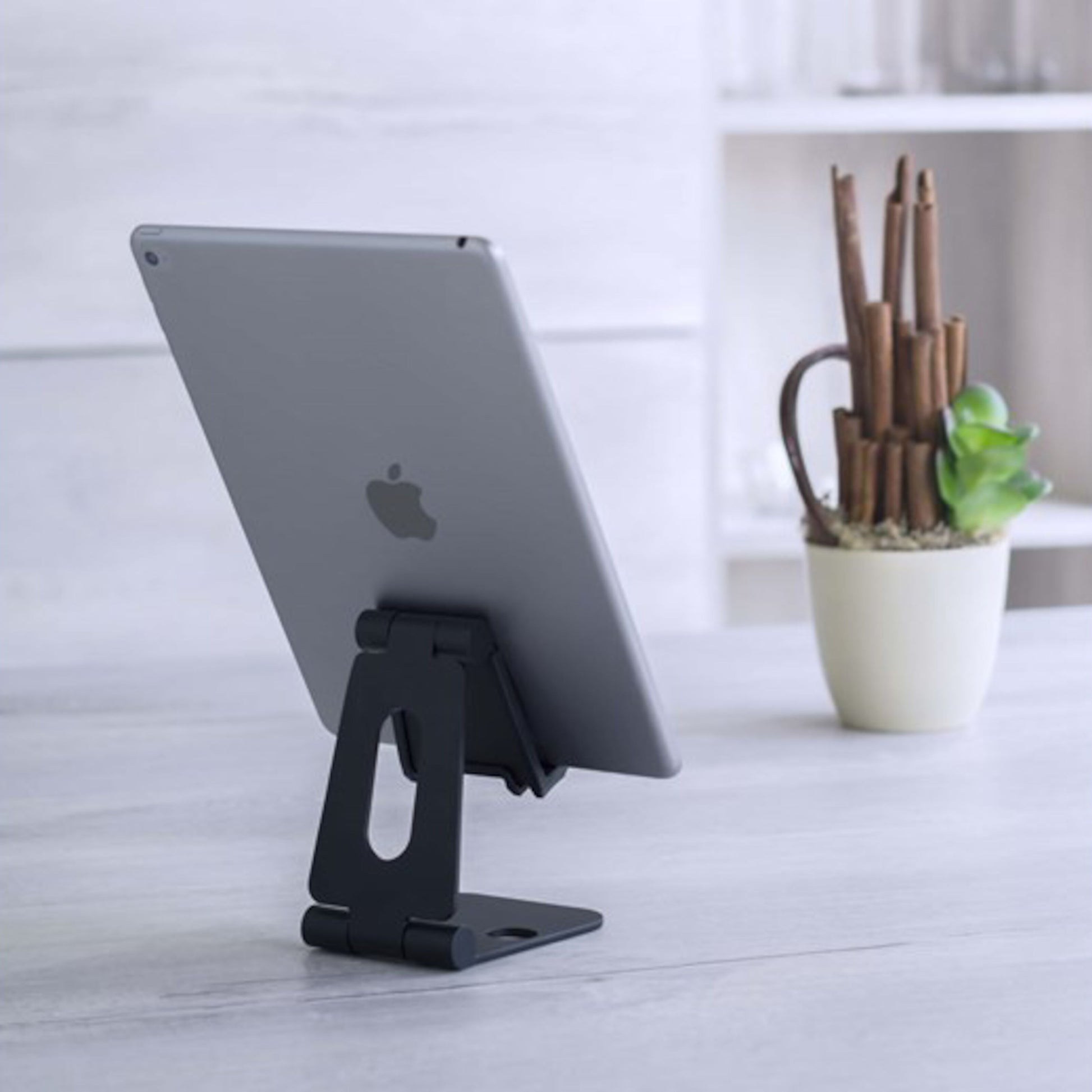 Stativ til mobil og tablet fra Linocell, holder tablet på lyst bord, grøn potteplante og hvide hylder synlige i baggrund.