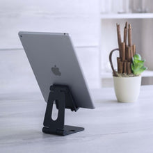 Stativ til mobil og tablet fra Linocell, holder tablet på lyst bord, grøn potteplante og hvide hylder synlige i baggrund.