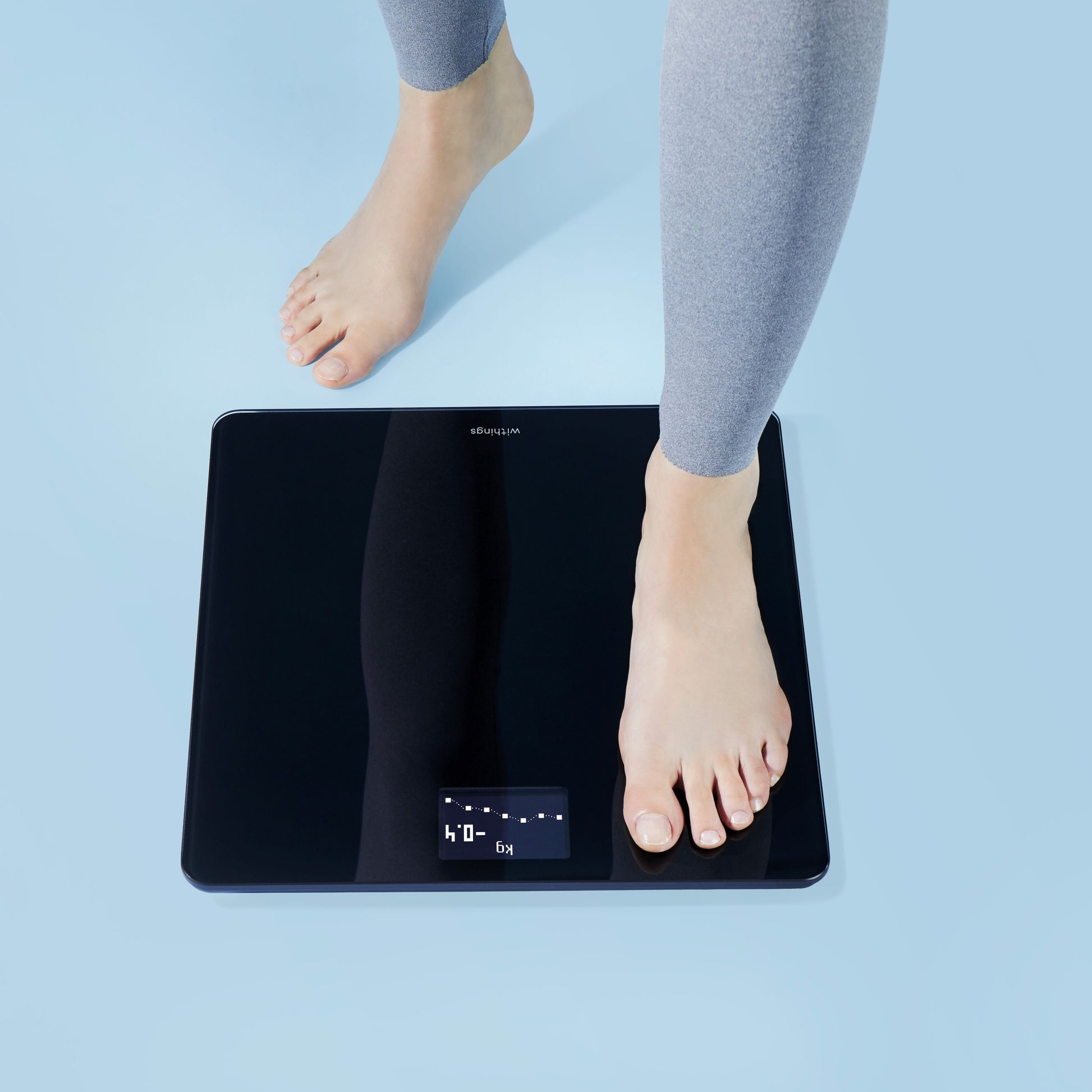Withings Krops personvægt med wifi Sort, sort overflade og digitalt display, vises brugt af person i grå leggings.