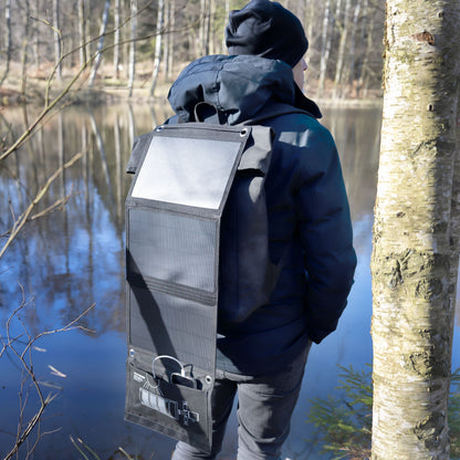 Rygsæk med Linocell Solcelleoplader 20 W monteret, sort jakke og hue, udendørs ved skovsø, set bagfra.
